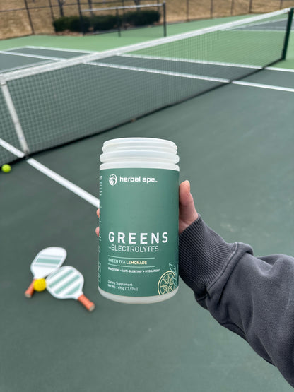 "A person holding a container of Herbal Ape Greens + Electrolytes (Green Tea Lemonade flavor) in front of a pickleball court. Two pickleball paddles and balls are placed on the court surface in the background. The supplement bottle has a matte green label with white and yellow text, emphasizing benefits like digestion, hydration, and anti-bloating.