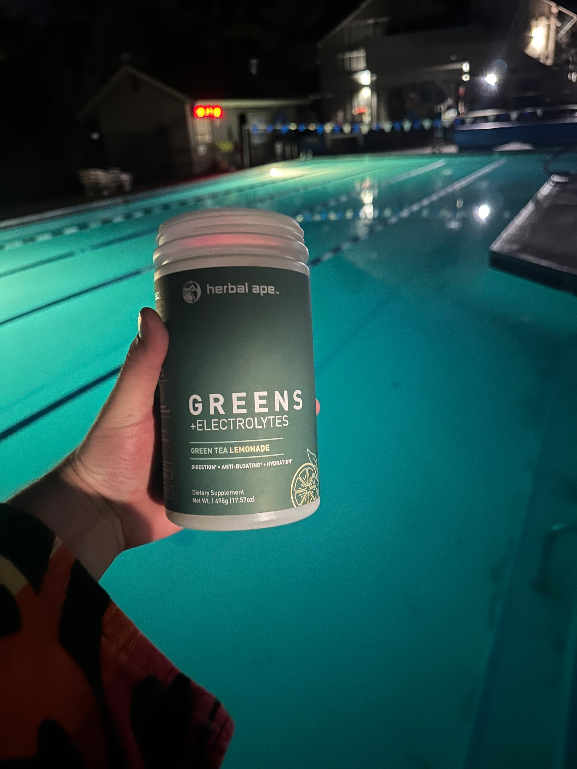 A hand holding a container of Herbal Ape Greens + Electrolytes (Green Tea Lemonade flavor) in front of a lit outdoor swimming pool at night. The label on the bottle highlights benefits such as digestion, hydration, and anti-bloating. The person appears to be wearing a multicolored towel.
