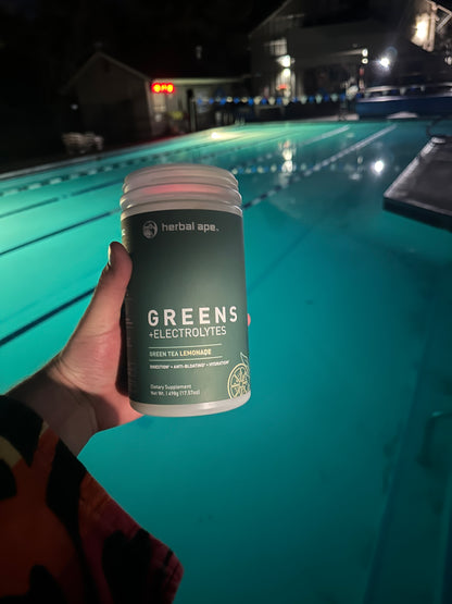A hand holding a container of Herbal Ape Greens + Electrolytes (Green Tea Lemonade flavor) in front of a lit outdoor swimming pool at night. The label on the bottle highlights benefits such as digestion, hydration, and anti-bloating. The person appears to be wearing a multicolored towel.